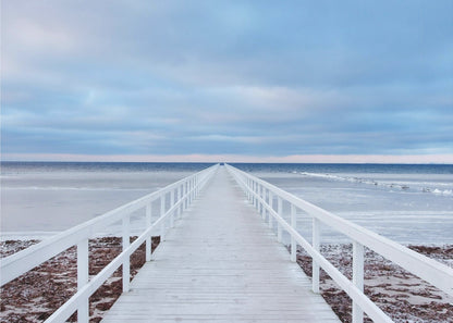 A framed photograph capturing a long, white wooden pier from a central perspective, leading the eye across a calm, icy sea towards the distant horizon under a cloudy blue sky. Print