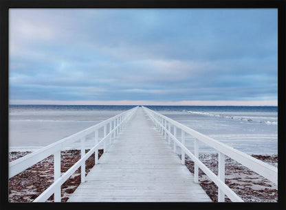 A framed photograph capturing a long, white wooden pier from a central perspective, leading the eye across a calm, icy sea towards the distant horizon under a cloudy blue sky. Print