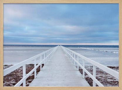 A framed photograph capturing a long, white wooden pier from a central perspective, leading the eye across a calm, icy sea towards the distant horizon under a cloudy blue sky. Print
