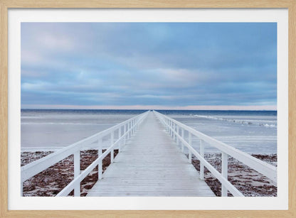 A framed photograph capturing a long, white wooden pier from a central perspective, leading the eye across a calm, icy sea towards the distant horizon under a cloudy blue sky. Print