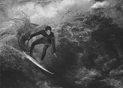 A dramatic black and white photograph of a surfer in a wetsuit skillfully riding down the face of a large, turbulent wave. The image is enclosed in a silver frame. Wall Art