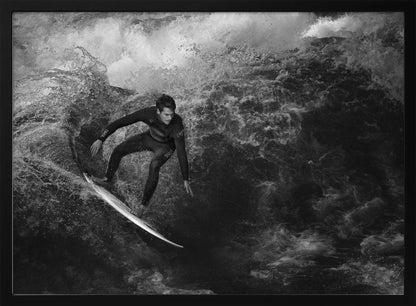 A dramatic black and white photograph of a surfer in a wetsuit skillfully riding down the face of a large, turbulent wave. The image is enclosed in a silver frame. Wall Art