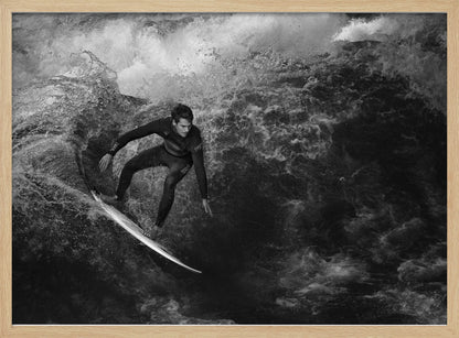 A dramatic black and white photograph of a surfer in a wetsuit skillfully riding down the face of a large, turbulent wave. The image is enclosed in a silver frame. Wall Art