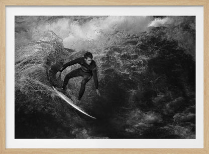 A dramatic black and white photograph of a surfer in a wetsuit skillfully riding down the face of a large, turbulent wave. The image is enclosed in a silver frame. Wall Art