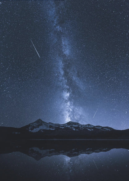 A breathtaking night photograph of the Milky Way galaxy stretching vertically across a deep blue, star-filled sky. Two shooting stars streak through the atmosphere above a dark, snow-dusted mountain range, which is perfectly reflected in the calm, mirror-like surface of a lake in the foreground. Artwork