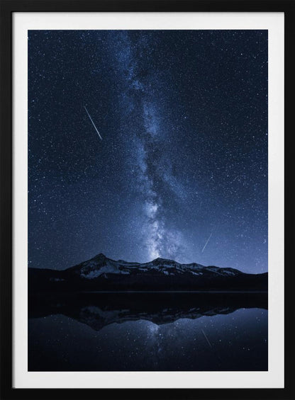 A breathtaking night photograph of the Milky Way galaxy stretching vertically across a deep blue, star-filled sky. Two shooting stars streak through the atmosphere above a dark, snow-dusted mountain range, which is perfectly reflected in the calm, mirror-like surface of a lake in the foreground. Artwork