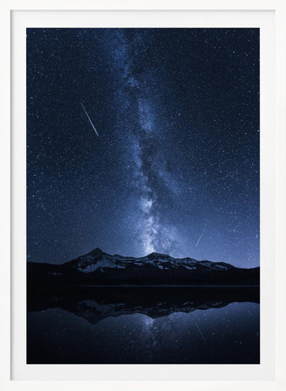 A breathtaking night photograph of the Milky Way galaxy stretching vertically across a deep blue, star-filled sky. Two shooting stars streak through the atmosphere above a dark, snow-dusted mountain range, which is perfectly reflected in the calm, mirror-like surface of a lake in the foreground. Artwork