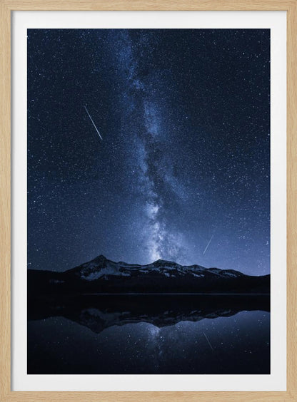 A breathtaking night photograph of the Milky Way galaxy stretching vertically across a deep blue, star-filled sky. Two shooting stars streak through the atmosphere above a dark, snow-dusted mountain range, which is perfectly reflected in the calm, mirror-like surface of a lake in the foreground. Artwork