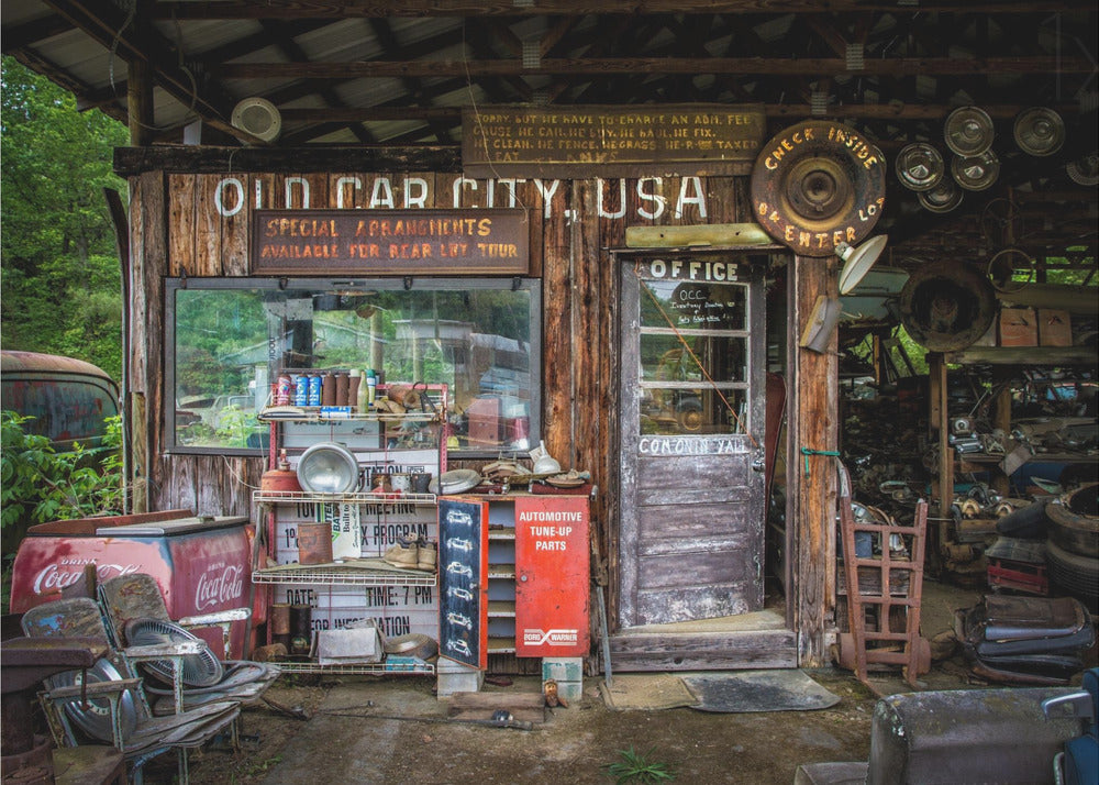 A framed photograph of the cluttered entrance to a rustic wooden building labeled 'Old Car City, USA'. The scene is filled with vintage memorabilia, including car parts, old signs, a red Coca-Cola cooler, and the weathered door to an office. Poster