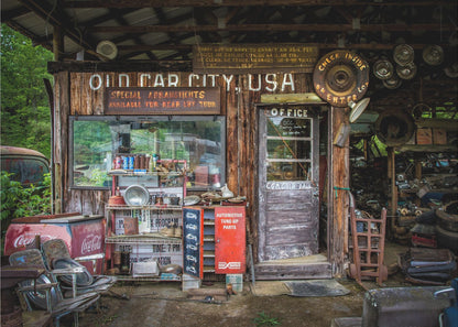A framed photograph of the cluttered entrance to a rustic wooden building labeled 'Old Car City, USA'. The scene is filled with vintage memorabilia, including car parts, old signs, a red Coca-Cola cooler, and the weathered door to an office. Poster