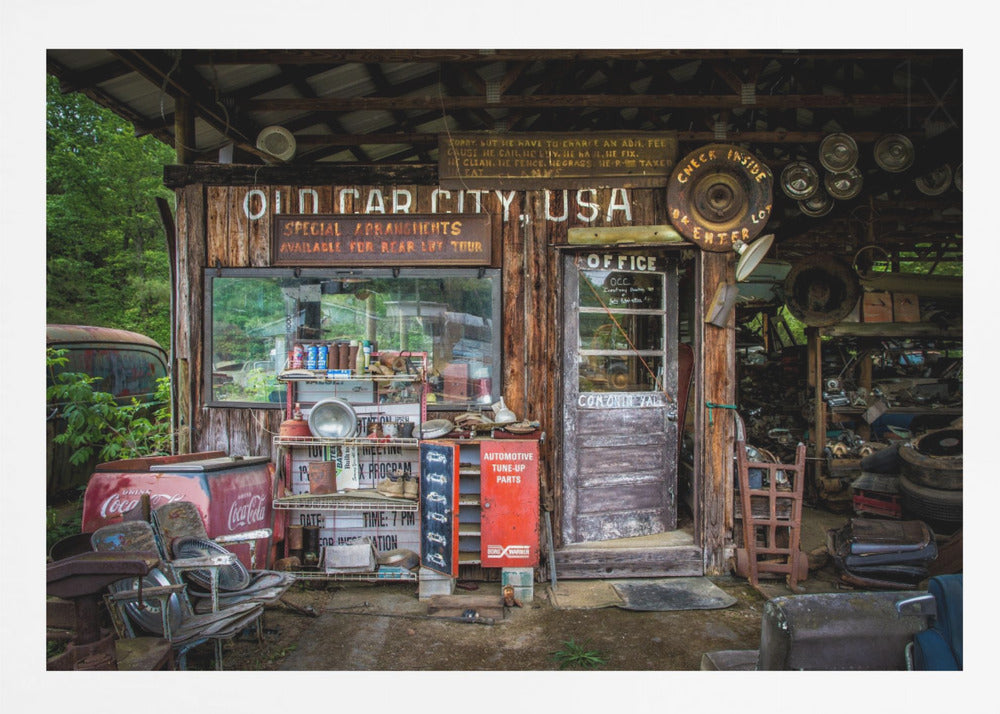 A framed photograph of the cluttered entrance to a rustic wooden building labeled 'Old Car City, USA'. The scene is filled with vintage memorabilia, including car parts, old signs, a red Coca-Cola cooler, and the weathered door to an office. Poster