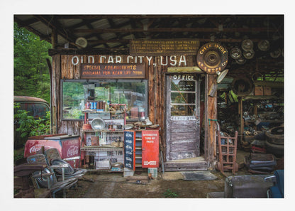 A framed photograph of the cluttered entrance to a rustic wooden building labeled 'Old Car City, USA'. The scene is filled with vintage memorabilia, including car parts, old signs, a red Coca-Cola cooler, and the weathered door to an office. Poster