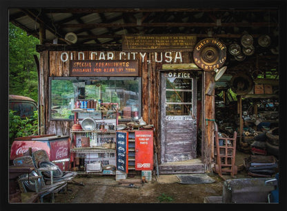A framed photograph of the cluttered entrance to a rustic wooden building labeled 'Old Car City, USA'. The scene is filled with vintage memorabilia, including car parts, old signs, a red Coca-Cola cooler, and the weathered door to an office. Poster