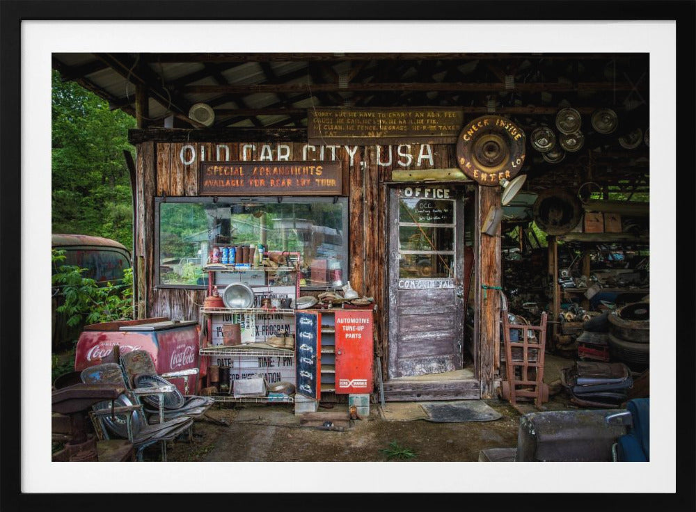A framed photograph of the cluttered entrance to a rustic wooden building labeled 'Old Car City, USA'. The scene is filled with vintage memorabilia, including car parts, old signs, a red Coca-Cola cooler, and the weathered door to an office. Poster
