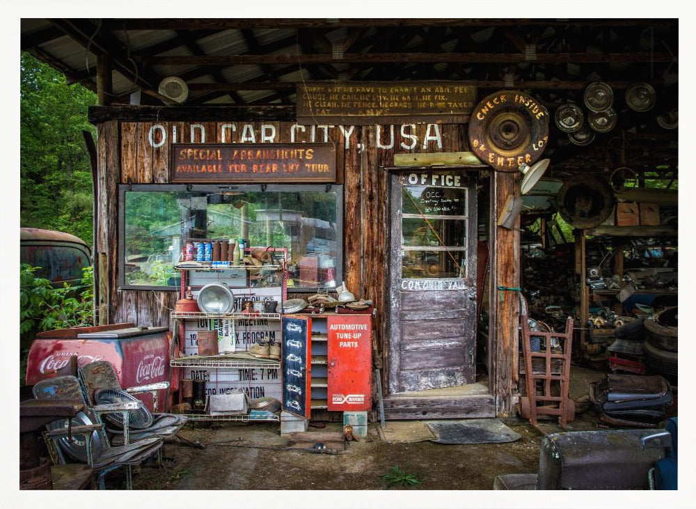 A framed photograph of the cluttered entrance to a rustic wooden building labeled 'Old Car City, USA'. The scene is filled with vintage memorabilia, including car parts, old signs, a red Coca-Cola cooler, and the weathered door to an office. Poster