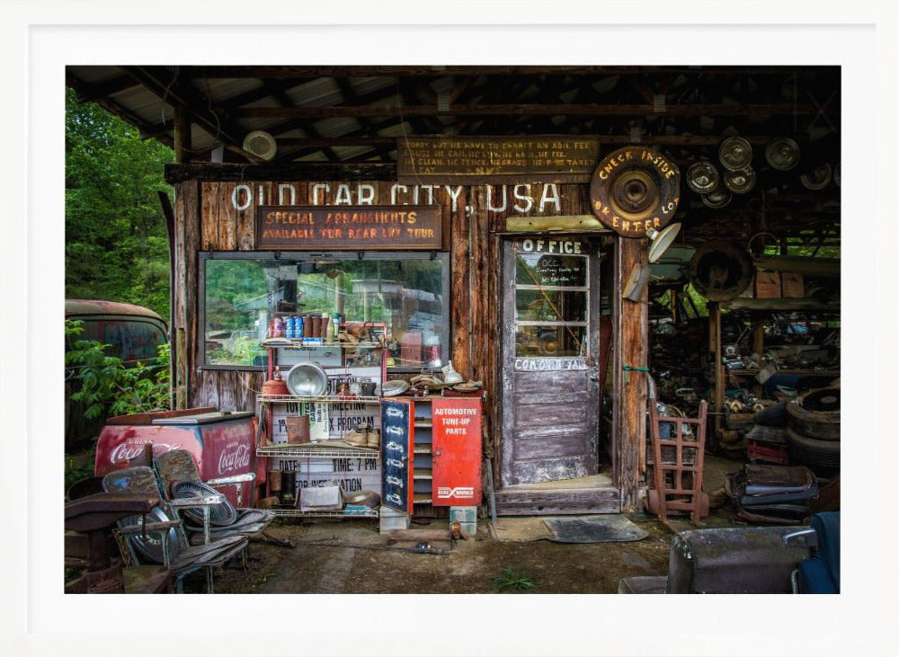 A framed photograph of the cluttered entrance to a rustic wooden building labeled 'Old Car City, USA'. The scene is filled with vintage memorabilia, including car parts, old signs, a red Coca-Cola cooler, and the weathered door to an office. Poster