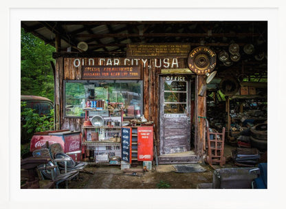 A framed photograph of the cluttered entrance to a rustic wooden building labeled 'Old Car City, USA'. The scene is filled with vintage memorabilia, including car parts, old signs, a red Coca-Cola cooler, and the weathered door to an office. Poster