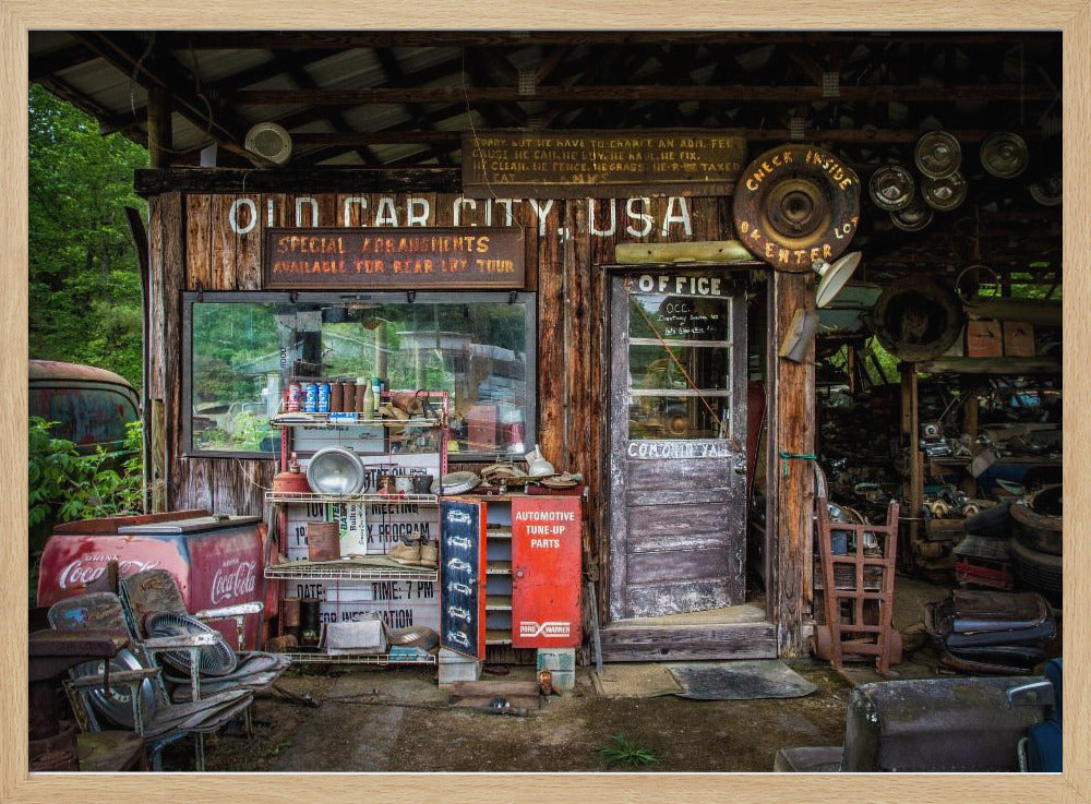 A framed photograph of the cluttered entrance to a rustic wooden building labeled 'Old Car City, USA'. The scene is filled with vintage memorabilia, including car parts, old signs, a red Coca-Cola cooler, and the weathered door to an office. Poster