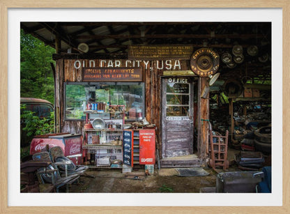 A framed photograph of the cluttered entrance to a rustic wooden building labeled 'Old Car City, USA'. The scene is filled with vintage memorabilia, including car parts, old signs, a red Coca-Cola cooler, and the weathered door to an office. Poster