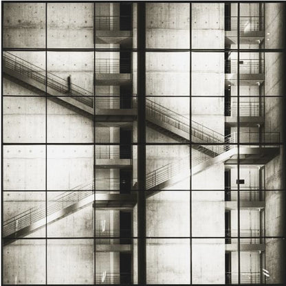 A framed black and white photograph of a modern concrete building's interior, seen through a grid of window panes. The image captures multiple levels of staircases and walkways crisscrossing in a complex, geometric pattern. A shadowy, blurred figure of a person can be seen walking up a staircase on the upper left. Wall Art