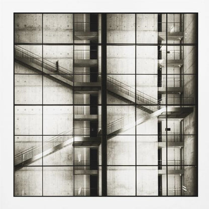 A framed black and white photograph of a modern concrete building's interior, seen through a grid of window panes. The image captures multiple levels of staircases and walkways crisscrossing in a complex, geometric pattern. A shadowy, blurred figure of a person can be seen walking up a staircase on the upper left. Wall Art