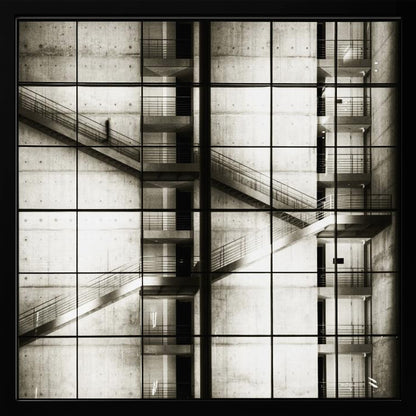 A framed black and white photograph of a modern concrete building's interior, seen through a grid of window panes. The image captures multiple levels of staircases and walkways crisscrossing in a complex, geometric pattern. A shadowy, blurred figure of a person can be seen walking up a staircase on the upper left. Wall Art