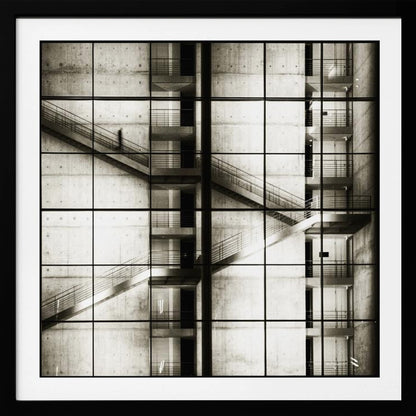 A framed black and white photograph of a modern concrete building's interior, seen through a grid of window panes. The image captures multiple levels of staircases and walkways crisscrossing in a complex, geometric pattern. A shadowy, blurred figure of a person can be seen walking up a staircase on the upper left. Wall Art