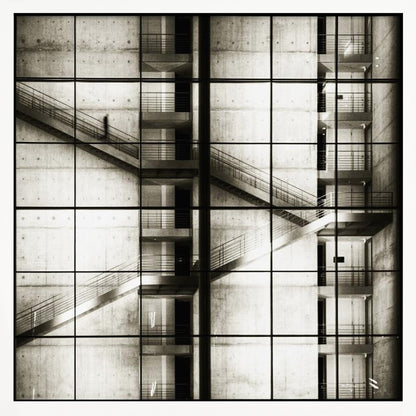 A framed black and white photograph of a modern concrete building's interior, seen through a grid of window panes. The image captures multiple levels of staircases and walkways crisscrossing in a complex, geometric pattern. A shadowy, blurred figure of a person can be seen walking up a staircase on the upper left. Wall Art