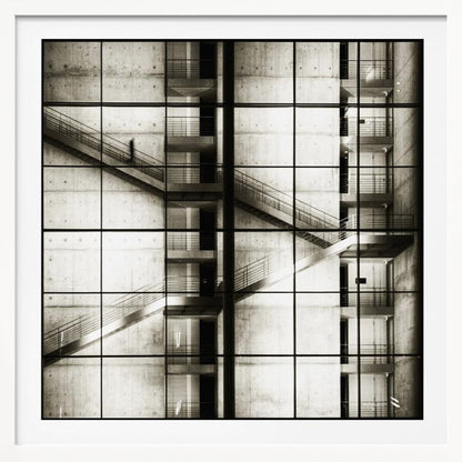 A framed black and white photograph of a modern concrete building's interior, seen through a grid of window panes. The image captures multiple levels of staircases and walkways crisscrossing in a complex, geometric pattern. A shadowy, blurred figure of a person can be seen walking up a staircase on the upper left. Wall Art