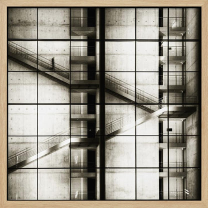A framed black and white photograph of a modern concrete building's interior, seen through a grid of window panes. The image captures multiple levels of staircases and walkways crisscrossing in a complex, geometric pattern. A shadowy, blurred figure of a person can be seen walking up a staircase on the upper left. Wall Art