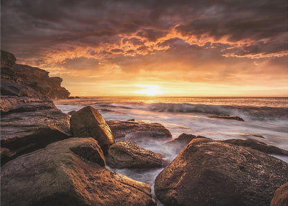 A framed photograph of a dramatic ocean sunset, with fiery orange clouds filling the sky. Large, dark rocks sit on the shore as long-exposure waves create a misty effect around them. Print