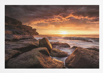 A framed photograph of a dramatic ocean sunset, with fiery orange clouds filling the sky. Large, dark rocks sit on the shore as long-exposure waves create a misty effect around them. Print