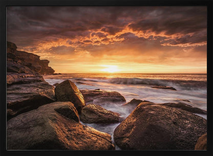 A framed photograph of a dramatic ocean sunset, with fiery orange clouds filling the sky. Large, dark rocks sit on the shore as long-exposure waves create a misty effect around them. Print