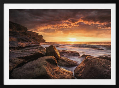 A framed photograph of a dramatic ocean sunset, with fiery orange clouds filling the sky. Large, dark rocks sit on the shore as long-exposure waves create a misty effect around them. Print
