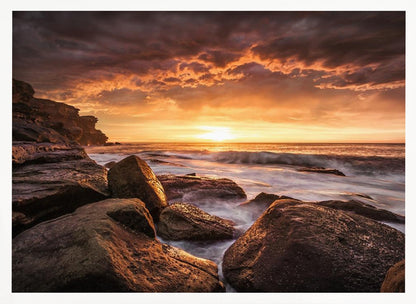 A framed photograph of a dramatic ocean sunset, with fiery orange clouds filling the sky. Large, dark rocks sit on the shore as long-exposure waves create a misty effect around them. Print