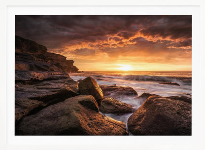 A framed photograph of a dramatic ocean sunset, with fiery orange clouds filling the sky. Large, dark rocks sit on the shore as long-exposure waves create a misty effect around them. Print