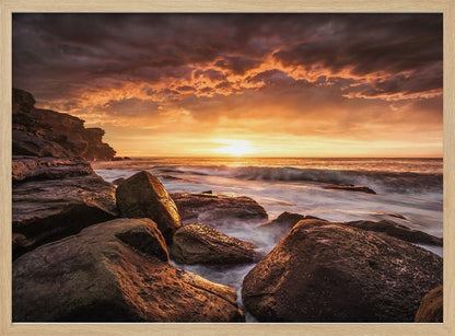 A framed photograph of a dramatic ocean sunset, with fiery orange clouds filling the sky. Large, dark rocks sit on the shore as long-exposure waves create a misty effect around them. Print