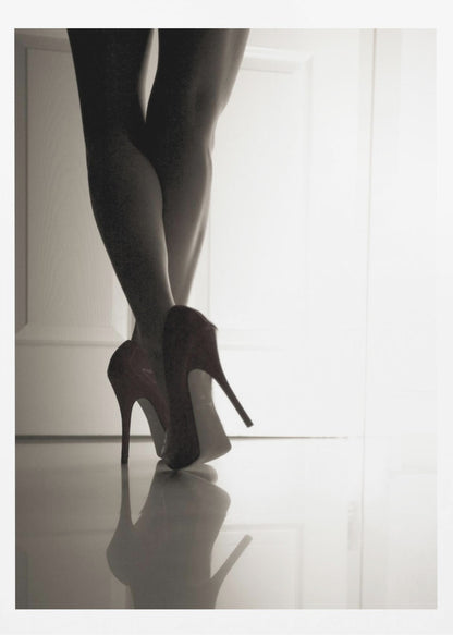 A low-angle, black and white photograph focusing on a woman's legs as she stands with them crossed. She wears dark red stiletto high heels, which cast a clear reflection on the glossy floor. Her legs are silhouetted against a bright white door in the background, creating a dramatic and sensual mood. Decor