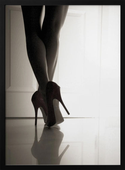 A low-angle, black and white photograph focusing on a woman's legs as she stands with them crossed. She wears dark red stiletto high heels, which cast a clear reflection on the glossy floor. Her legs are silhouetted against a bright white door in the background, creating a dramatic and sensual mood. Decor