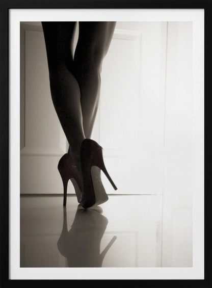 A low-angle, black and white photograph focusing on a woman's legs as she stands with them crossed. She wears dark red stiletto high heels, which cast a clear reflection on the glossy floor. Her legs are silhouetted against a bright white door in the background, creating a dramatic and sensual mood. Decor