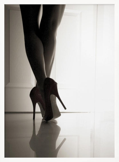 A low-angle, black and white photograph focusing on a woman's legs as she stands with them crossed. She wears dark red stiletto high heels, which cast a clear reflection on the glossy floor. Her legs are silhouetted against a bright white door in the background, creating a dramatic and sensual mood. Decor