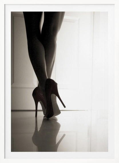 A low-angle, black and white photograph focusing on a woman's legs as she stands with them crossed. She wears dark red stiletto high heels, which cast a clear reflection on the glossy floor. Her legs are silhouetted against a bright white door in the background, creating a dramatic and sensual mood. Decor