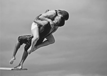 A framed, black and white photograph capturing a dynamic and romantic moment. A man is leaping off a diving board, carrying a woman who is wrapped around him in a passionate embrace, their faces close together. The background is a simple, plain gray, emphasizing their mid-air pose. Wall Art
