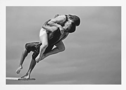 A framed, black and white photograph capturing a dynamic and romantic moment. A man is leaping off a diving board, carrying a woman who is wrapped around him in a passionate embrace, their faces close together. The background is a simple, plain gray, emphasizing their mid-air pose. Wall Art