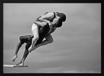 A framed, black and white photograph capturing a dynamic and romantic moment. A man is leaping off a diving board, carrying a woman who is wrapped around him in a passionate embrace, their faces close together. The background is a simple, plain gray, emphasizing their mid-air pose. Wall Art