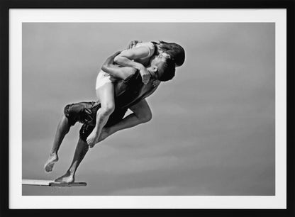 A framed, black and white photograph capturing a dynamic and romantic moment. A man is leaping off a diving board, carrying a woman who is wrapped around him in a passionate embrace, their faces close together. The background is a simple, plain gray, emphasizing their mid-air pose. Wall Art