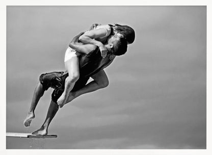 A framed, black and white photograph capturing a dynamic and romantic moment. A man is leaping off a diving board, carrying a woman who is wrapped around him in a passionate embrace, their faces close together. The background is a simple, plain gray, emphasizing their mid-air pose. Wall Art
