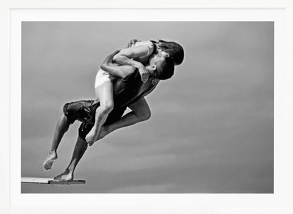 A framed, black and white photograph capturing a dynamic and romantic moment. A man is leaping off a diving board, carrying a woman who is wrapped around him in a passionate embrace, their faces close together. The background is a simple, plain gray, emphasizing their mid-air pose. Wall Art