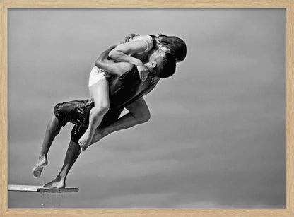 A framed, black and white photograph capturing a dynamic and romantic moment. A man is leaping off a diving board, carrying a woman who is wrapped around him in a passionate embrace, their faces close together. The background is a simple, plain gray, emphasizing their mid-air pose. Wall Art