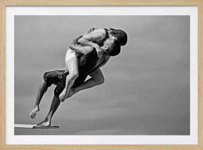A framed, black and white photograph capturing a dynamic and romantic moment. A man is leaping off a diving board, carrying a woman who is wrapped around him in a passionate embrace, their faces close together. The background is a simple, plain gray, emphasizing their mid-air pose. Wall Art